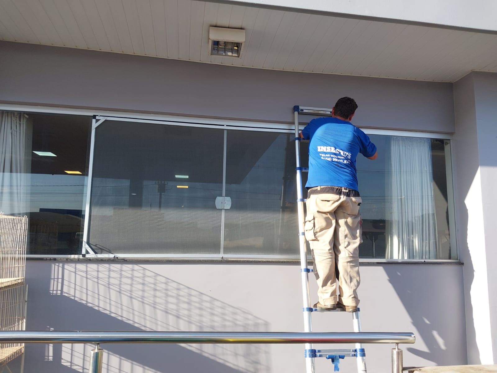 Instalador da Insectus em escada fixando tela mosquiteira em janela ampla de estabelecimento comercial