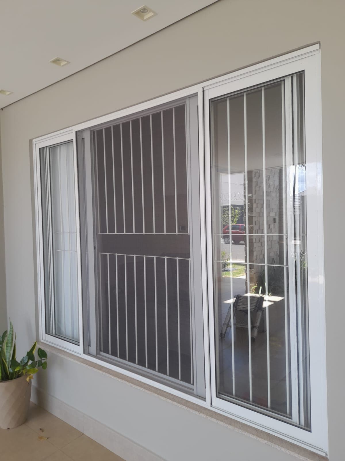 Janela com grade de proteção e tela mosquiteira instalada em varanda residencial, com vaso de planta