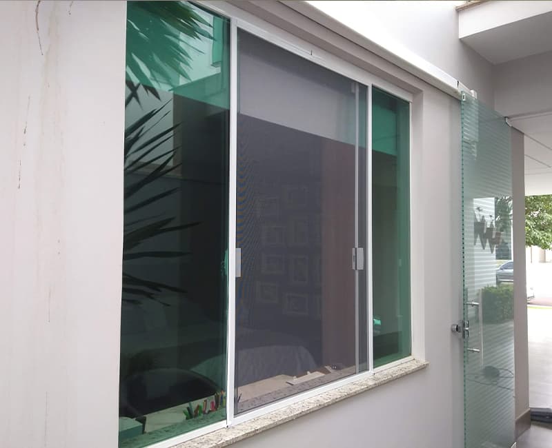 Janela blindex de vidro verde com tela mosquiteira de correr em perfil branco instalada na própria abertura, fachada residencial