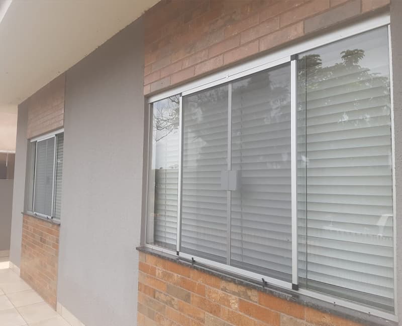 Janelas blindex com tela mosquiteira de perfil prata em fachada de tijolo aparente, com persiana fechada do lado de dentro